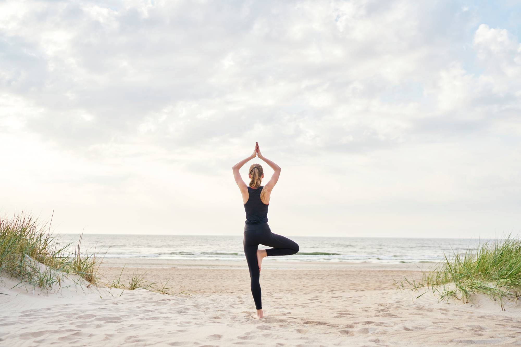 A woman in black yoga suit doing tree pose on the beach.